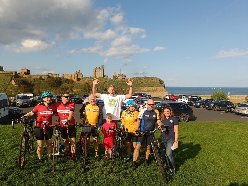 A group of cyclists and their supporters stand together, with a tower and the sea beyond