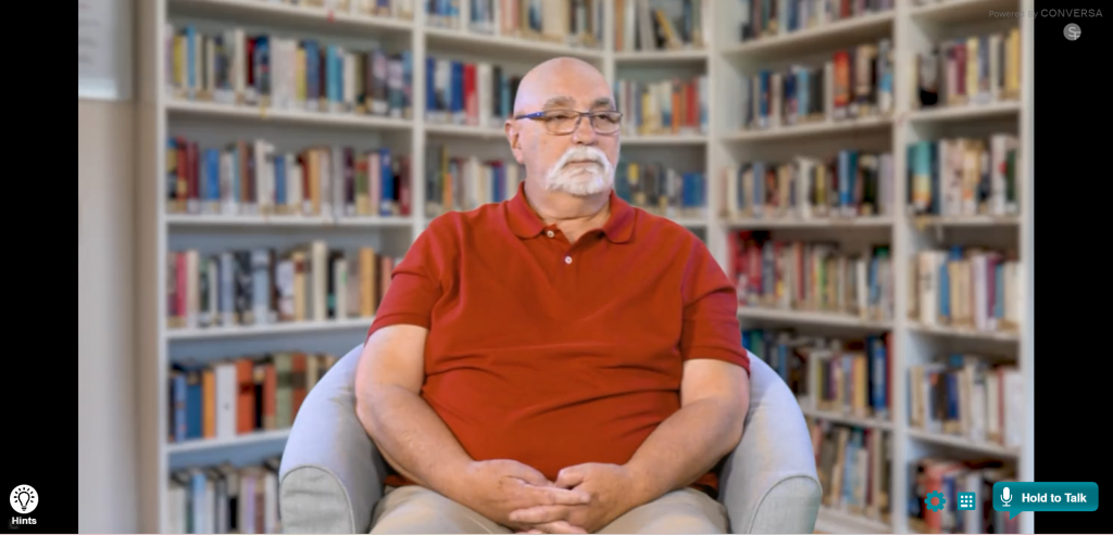 AI Pete is an older man, sitting in a comfortable chair in front of a full book case. He is casually dressed wearing a dark orange, short-sleeved polo shirt with light-coloured trousers. He has a short, silver, well-trimmed beard and is wearing glasses. 